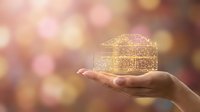Golden wireframe house model floating above open palm with warm bokeh background representing real estate investment and property dreams.