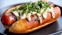 Grilled hot dog in toasted brioche bun topped with melted cheese sauce, fresh chopped herbs and microgreens served on dark plate, close up view.