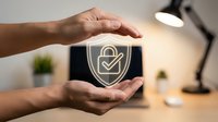 Hands protecting digital security shield with checkmark symbol representing cybersecurity protection and data safety in modern office environment.