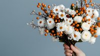 Minimalist floral arrangement with white peonies and orange berries held against gray background, showcasing modern botanical styling for wedding and events.