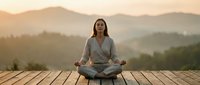 Young woman practicing yoga meditation on wooden deck at sunrise with mountain landscape background for wellness and mindfulness lifestyle content.