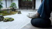 Person meditating in traditional Japanese zen garden with raked gravel patterns and bonsai trees creating peaceful mindfulness atmosphere for relaxation.