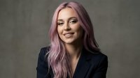 Young professional woman with pink purple hair smiling confidently in black blazer against textured gray background for business portraits.