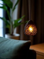 Cozy pendant lamp with woven rattan shade casting warm glow over green cushion, creating intimate atmosphere with blurred houseplant in background.