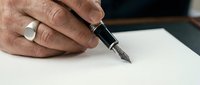 Mature male hand with silver ring holding fountain pen over blank white paper, ready to write or sign a document.