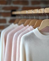 Soft pastel sweaters hanging on wooden hangers against brick wall background, featuring gentle gradient from white to pink tones, showcasing minimalist wardrobe organization.