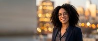 Young woman with dark curly hair and glasses wearing black blazer against blurred city lights at dusk, suitable for corporate, business, and professional lifestyle content.