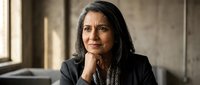 Professional mature South Asian businesswoman in black suit sitting thoughtfully by window in modern office environment for corporate presentations.