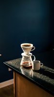 Pour over coffee setup with ceramic dripper, glass carafe, and white mug on dark granite countertop against moody blue-black background. Specialty coffee brewing method for cafes and home use.