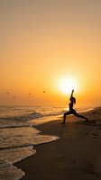 Woman practicing yoga warrior pose on beach at golden sunset with ocean waves and flying birds creating peaceful meditation scene.