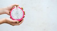 Woman's hands holding halved dragon fruit with white flesh and small black seeds against neutral background, showcasing fresh tropical produce for food and nutrition content.