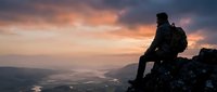 Silhouetted hiker with backpack sitting on mountain peak at sunset, overlooking vast valley landscape below with dramatic golden and purple sky.