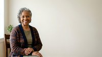 Senior African American woman with gray hair wearing burgundy cardigan and olive top, smiling at camera in bright interior space with plant and neutral wall background, suitable for corporate, healthcare, and lifestyle editorial use.