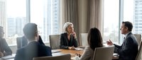 Diverse business team in formal meeting discussing strategy and collaboration in modern high-rise office with city skyline view through windows.