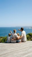 Family of four enjoying coastal ocean view from wooden deck, parents with two young children sitting together pointing at seascape, summer vacation and bonding moment.