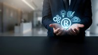 Professional businessman in dark suit holding glowing trademark symbol with innovation icons including lightbulb, gears and brain in modern office setting.