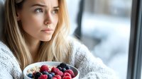 Young blonde woman in cozy sweater enjoying healthy breakfast bowl with fresh berries and granola by window, morning wellness lifestyle concept.
