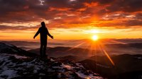 Person with arms outstretched standing on mountain peak at sunset, silhouetted against golden sky with dramatic clouds and sun rays over distant valleys.