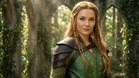 Young woman with long reddish-blonde braided hair wearing medieval fantasy warrior costume with green tunic and brown leather armor, standing in enchanted forest setting with hanging vines and natural sunlight filtering through trees.