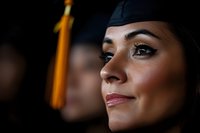 Close-up portrait of Hispanic female graduate in academic cap with gold tassel, showing confident smile and professional makeup against dark background.