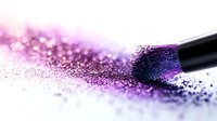 Scattered purple and pink glitter makeup powder with professional cosmetic brush on white background, macro close-up shot showing sparkly texture and shimmer effect.