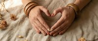 Hands of a woman wearing boho bracelets forming a heart shape on linen fabric with dried flowers, symbolizing love and mindfulness.