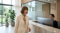 Professional businesswoman in beige suit walking through modern office lobby with reception desk. Corporate executive in contemporary workspace interior.