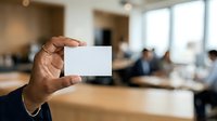Businessman holding blank white business card in modern office environment with colleagues working in background.