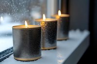 Glowing candles in metallic holders on windowsill against rainy window create cozy atmosphere. Soft focus on burning flames brings warmth to moody winter scene.
