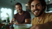 Young bearded man in yellow t-shirt smiling at dinner table with friend in background. Natural lifestyle scene with warm lighting and shallow depth of field.