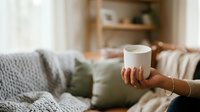 Cropped view of woman holding white mug in cozy living room with knit blanket, pillows and wooden shelving in background. Perfect for beverage, lifestyle and home decor marketing.
