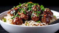 Glazed meatballs with sesame seeds and fresh herbs served over noodles in white bowl. Asian fusion cuisine for restaurant menus and food blogs.