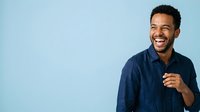 Young man with dark skin tone and black hair wearing navy blue casual shirt, smiling and pointing at camera against light blue background for advertising and promotional use.