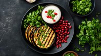 Grilled eggplant slices with pomegranate seeds, yogurt sauce and fresh herbs on black plate. Mediterranean cuisine served on dark stone background.