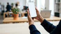 Person holding blank smartphone with white screen in modern office workspace, suitable for app mockups and digital marketing presentations.