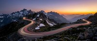 Winding mountain road with light trails at sunset creating dramatic curves through alpine landscape with snow-capped peaks and colorful sky.
