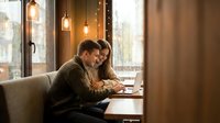 Young couple working together on laptop in cozy cafe with warm lighting and pendant lamps creating intimate atmosphere for remote work or study session.