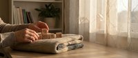 Woman hands placing wooden blocks on folded knitted sweaters in cozy home interior with natural sunlight streaming through window curtains.