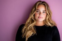 Young Caucasian woman with wavy blonde hair and natural makeup wearing black sweater against purple background, soft lighting creates gentle shadows and depth.