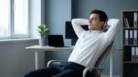 Young businessman relaxing at desk with hands behind head in modern office workspace taking break from laptop work.