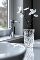 White magnolia flowers in crystal vase on bathroom sink counter with chrome faucet, modern minimalist interior design with natural window light and reflections.