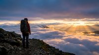 Hiker with backpack standing on mountain peak above clouds at golden sunset, adventure and travel concept.