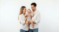Happy young Caucasian family with baby boy in striped outfit standing together on white background, parents smiling at child in casual clothing.