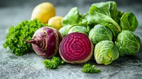 Fresh organic vegetables including sliced beetroot showing vibrant purple interior, brussels sprouts, lemons and curly parsley on rustic gray background. Food photography.