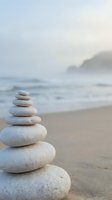 Zen stone stack on peaceful beach with soft waves and misty coastline creating tranquil meditation atmosphere for wellness and mindfulness practices.