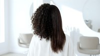 Young mixed-race woman with curly and straight hair from behind in white robe at modern bathroom mirror. Morning skincare and beauty routine concept.