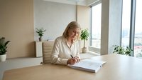 Professional blonde businesswoman in white blazer writing documents at modern office desk near window with city view and green plants.