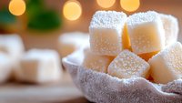 Homemade white marshmallow cubes dusted with powdered sugar in rustic linen napkin, warm bokeh lights background creates cozy atmosphere.