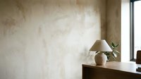 Minimalist interior with textured beige wall, ceramic table lamp and plant on wooden desk near window creating warm natural lighting atmosphere.