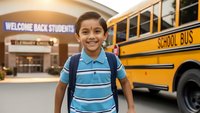 Happy Hispanic elementary school boy with backpack standing in front of yellow school bus and school building on first day back to school.